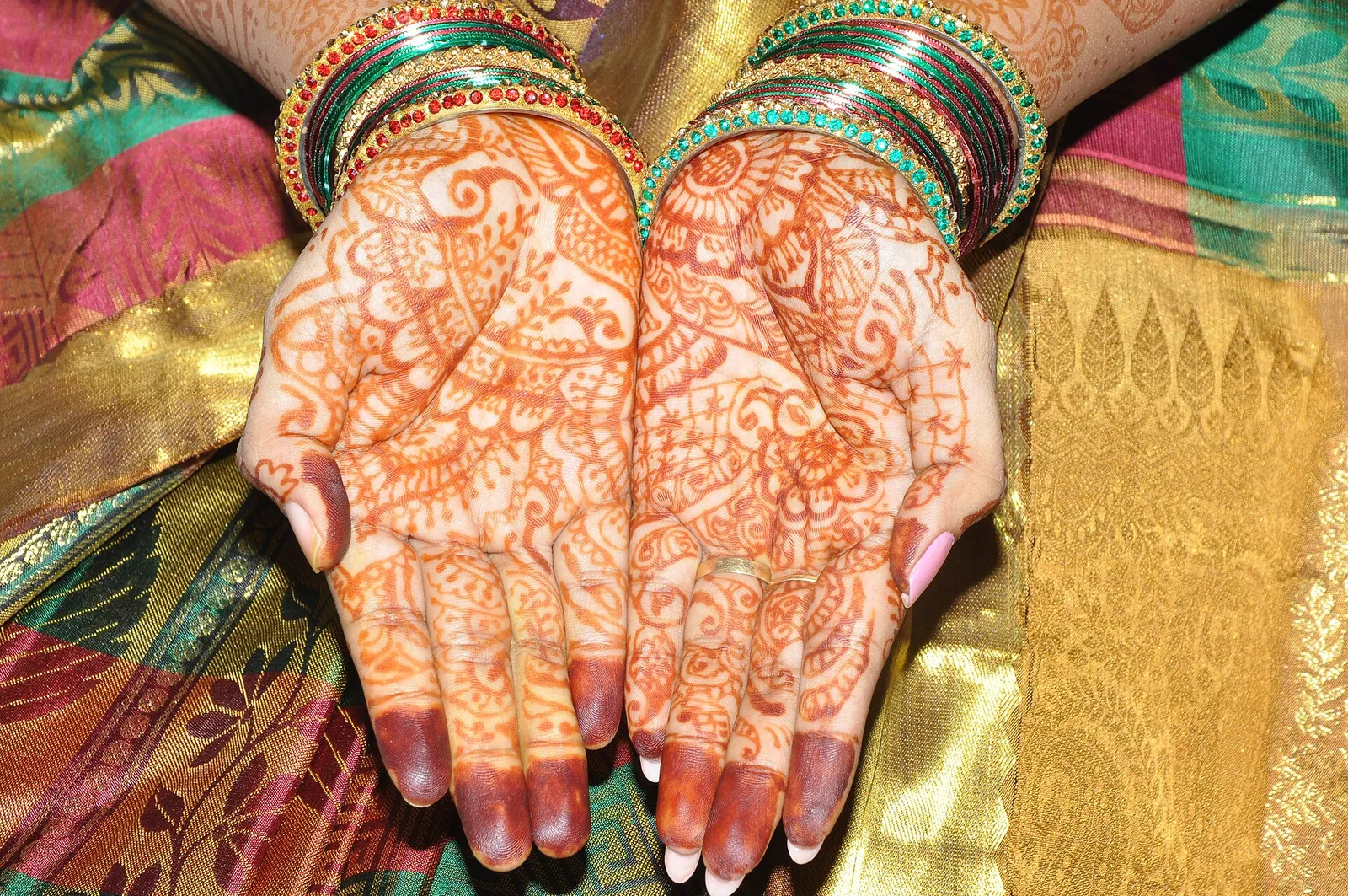 A bride’s hands with fine henna patterns and green bangles, resting on a silk saree with a zari border