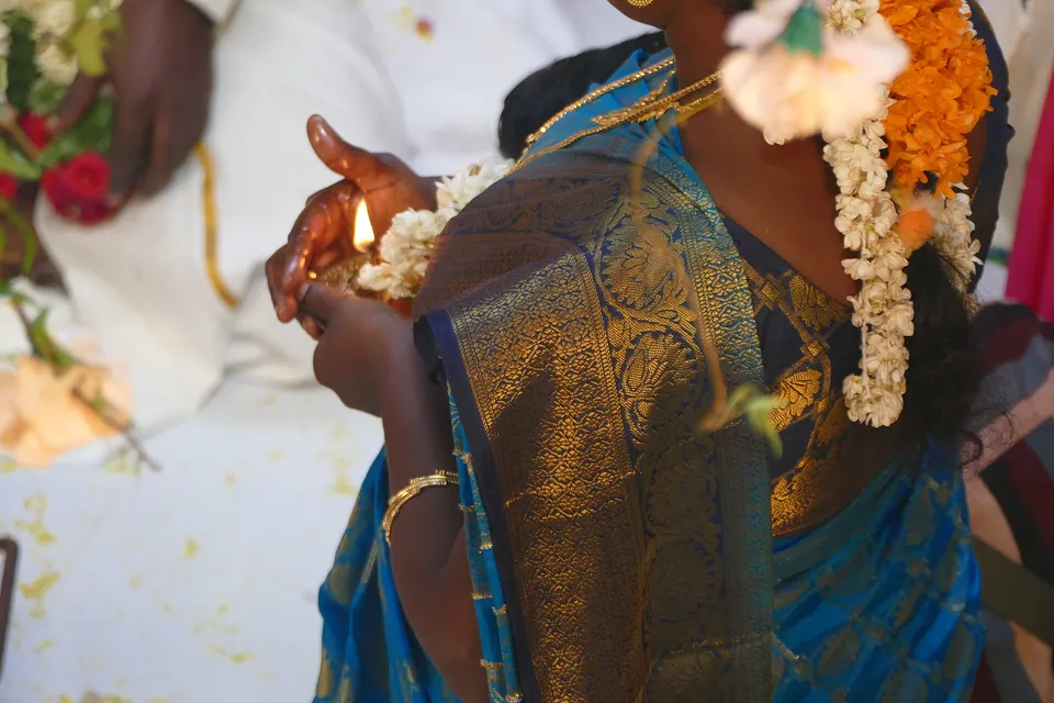 Hands holding a lit brass lamp beside a silk saree and jasmine flowers