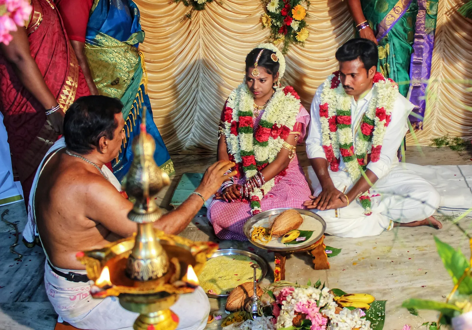 A bride and groom in flower garlands seated before the sacred fire as a priest performs the wedding rites