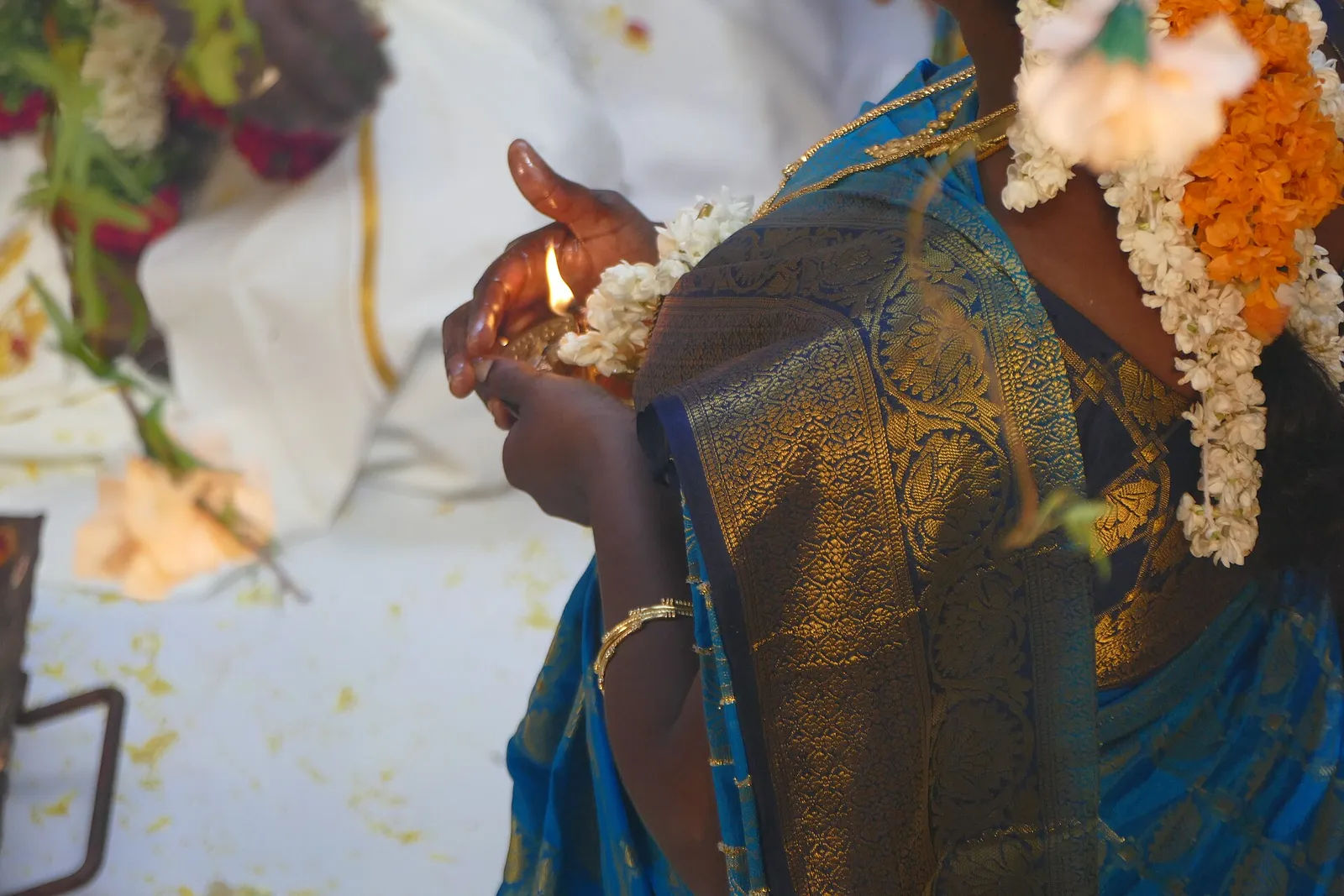 A woman in a blue silk saree holding a lit brass lamp, with jasmine in her hair