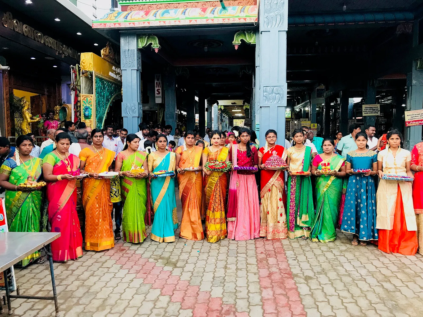 Women in bright silk sarees stand in a line holding decorated trays to welcome the groom’s party